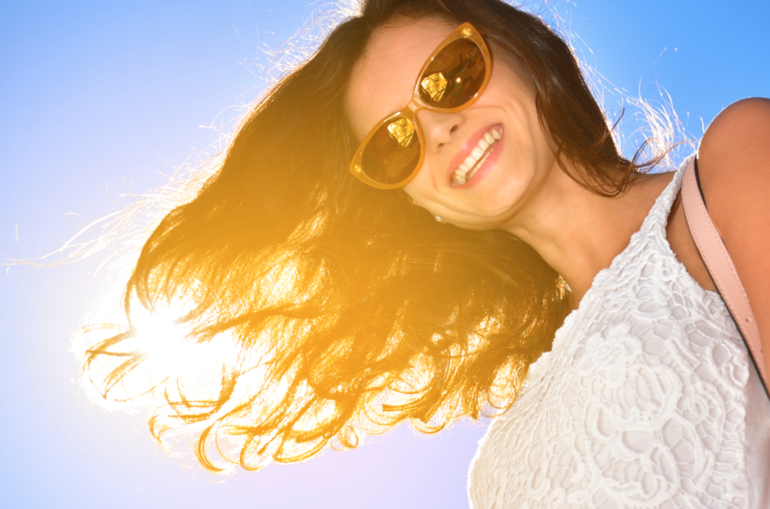 Women with sunglasses and wavy hair smiling in front of the sun.